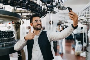 This image shows a manufacturer on a video call using his mobile phone. He’s standing in front of his equipment and giving the person he’s talking with a thumbs up. 