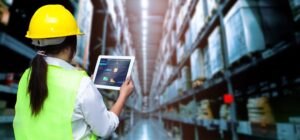 A woman in a high visibility vest types on a laptop in an industrial setting.