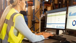 This image shows a female factory worker at a computer. She’s wearing a hard hat and a yellow vest while typing. A monitor on her right shows various charts and graphs. 